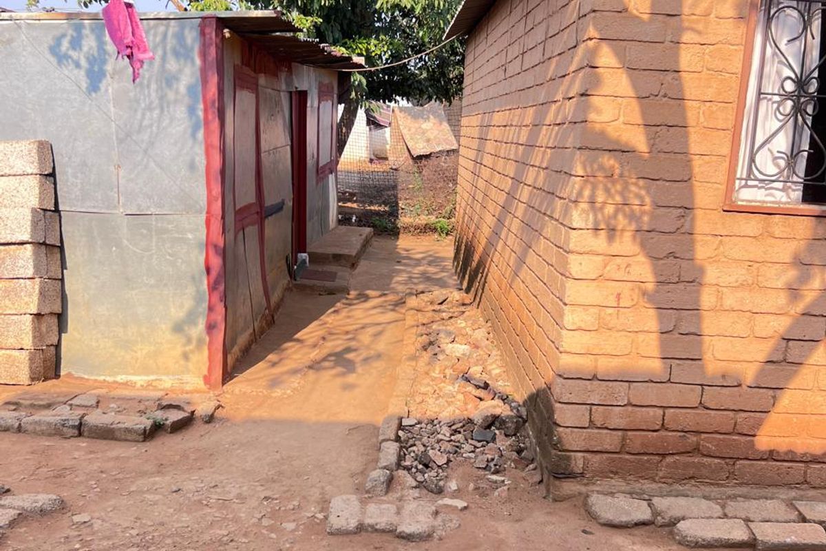 Exterior view showing a brick structure and a secondary corrugated metal outbuilding with an unpaved dirt pathway between them.