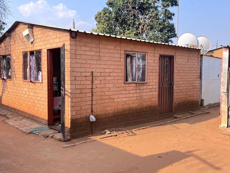 Exterior view of a single-story brick structure with metal roofing, security gates, and metal window frames, situated on an unpaved lot.