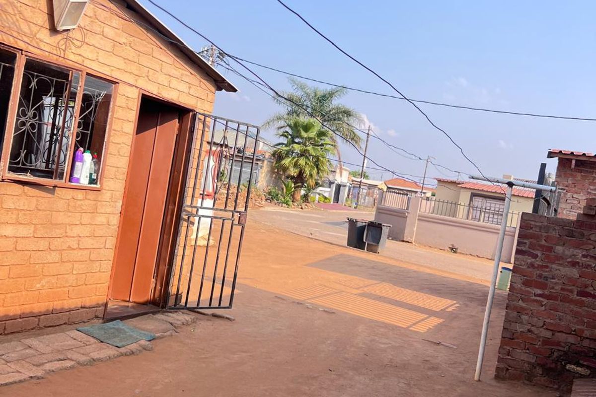 Exterior view of a brick house featuring a security gate and bars, with an unpaved driveway area.