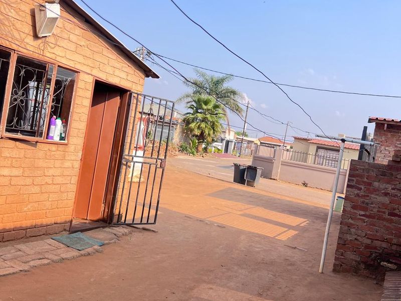 Exterior view of a brick house featuring a security gate and bars, with an unpaved driveway area.