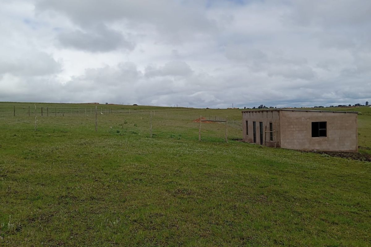 A view of a large vacant residential plot featuring grassy terrain, wire fencing, and a small, unfinished masonry structure.