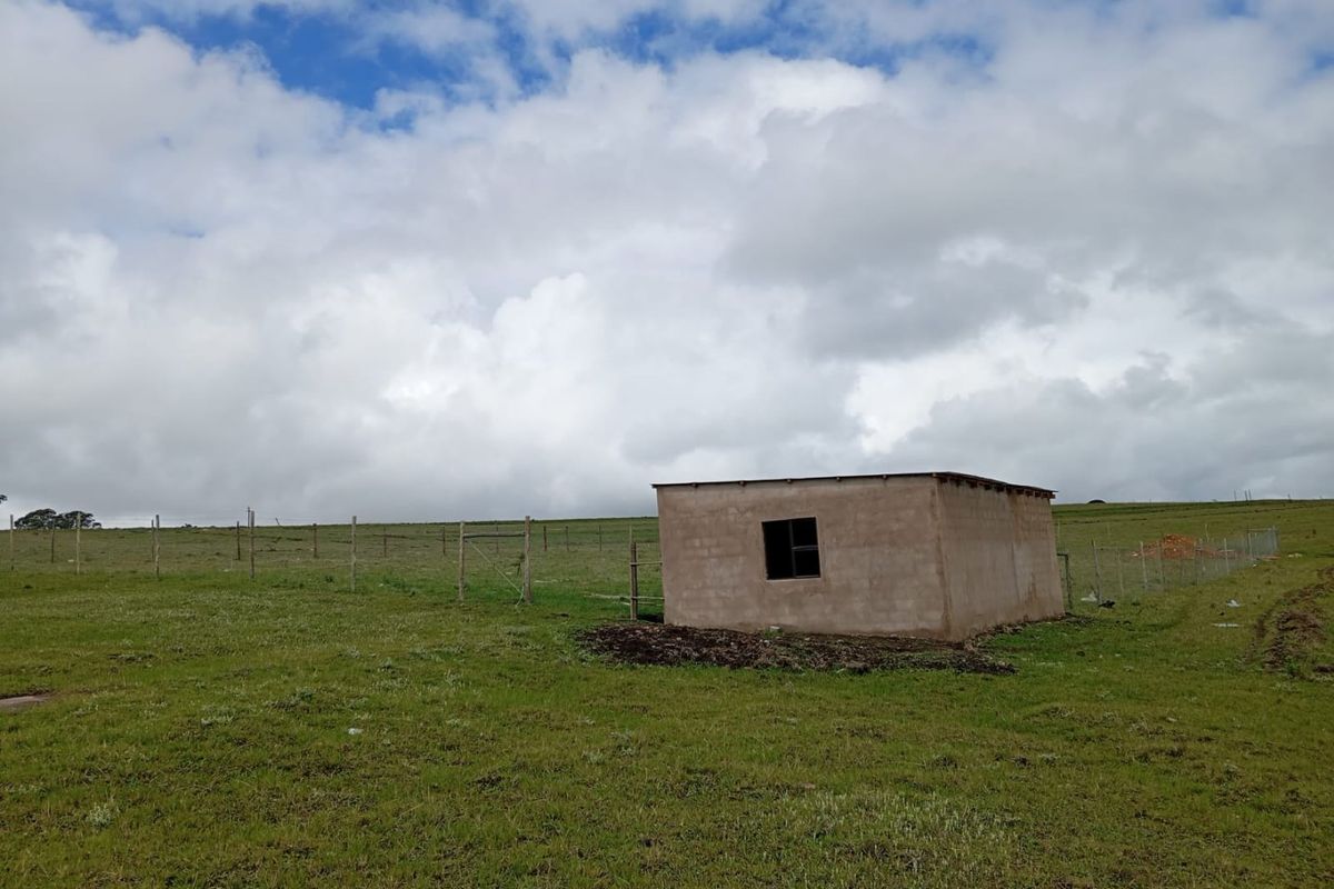 A small, unfinished masonry structure situated on a large, open plot of vacant land enclosed by wire fencing.