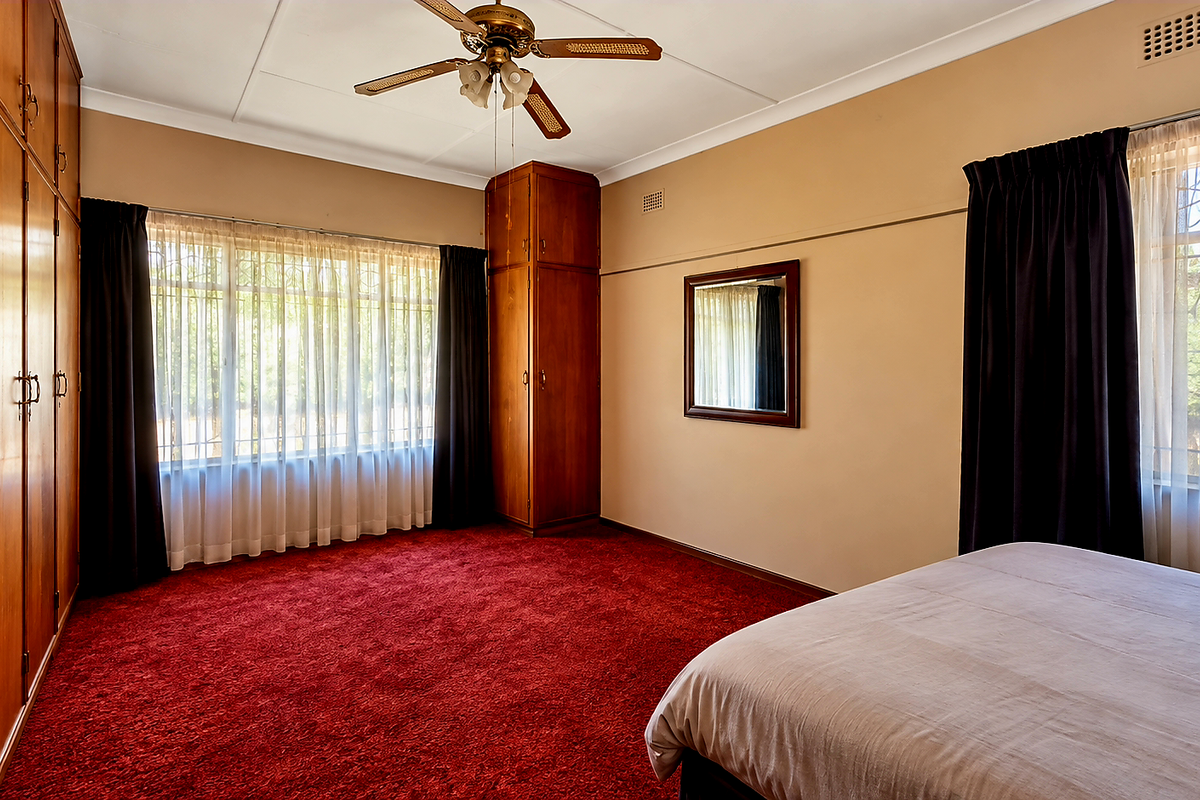 Spacious bedroom featuring extensive built-in wooden cabinetry, ceiling fan, and large windows with security bars, finished with wall-to-wall carpeting.