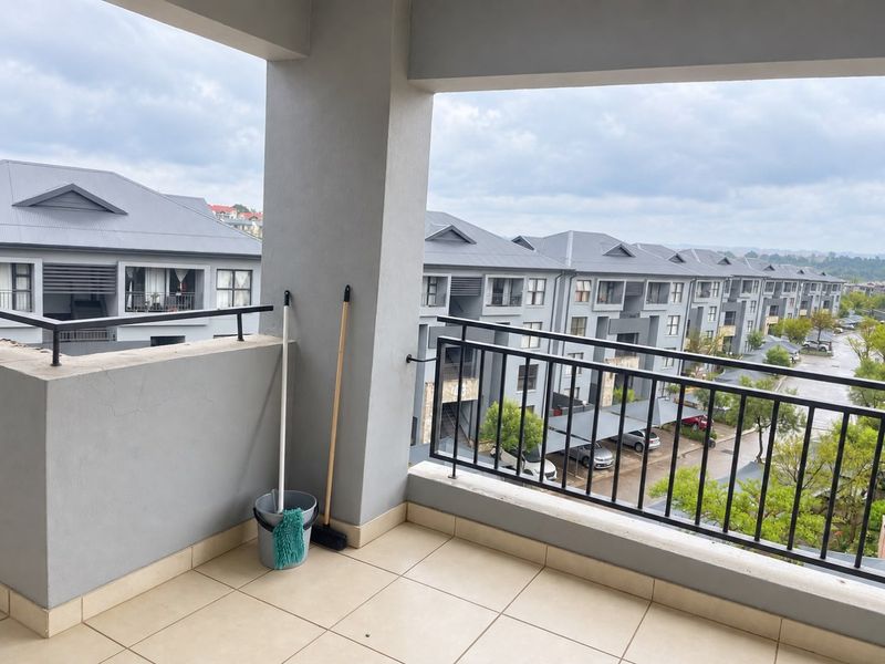 A spacious covered balcony featuring tiled flooring and a metal balustrade, offering an elevated view of the surrounding apartment complex.