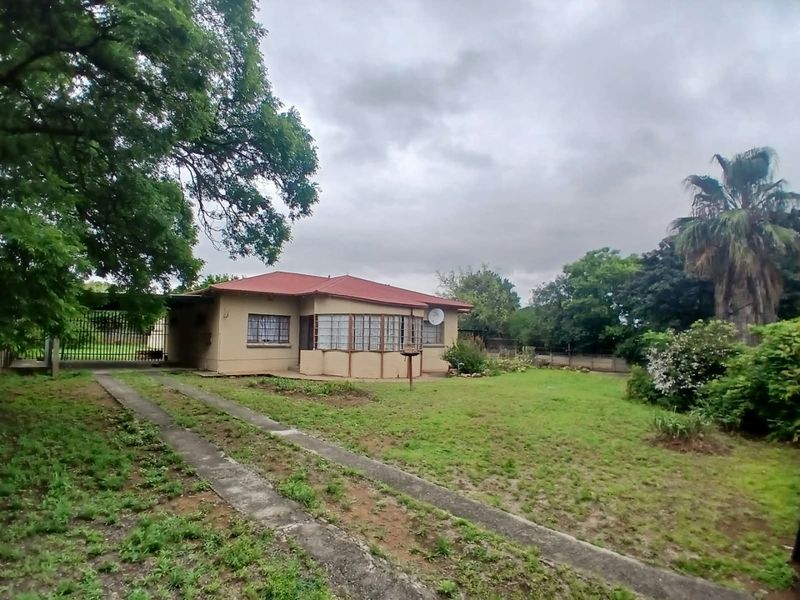 Front view of a single-story house featuring a metal roof, concrete driveway, and established garden with perimeter fencing.