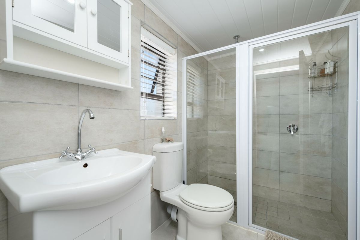 A clean, modern bathroom featuring a white vanity, glass-enclosed shower, neutral wall tiling, and a functional toilet.