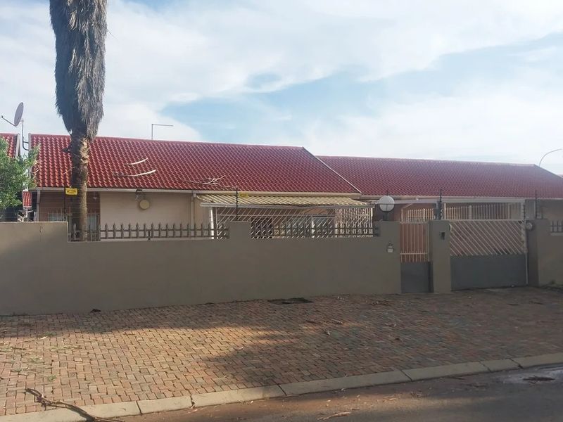 Front exterior view of a single-story house featuring a red tiled roof, perimeter wall with security fencing, and a paved driveway.