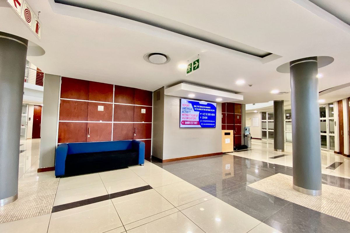 A spacious commercial building lobby featuring polished tiled flooring, structural columns, recessed ceiling lighting, and clear emergency exit signage.