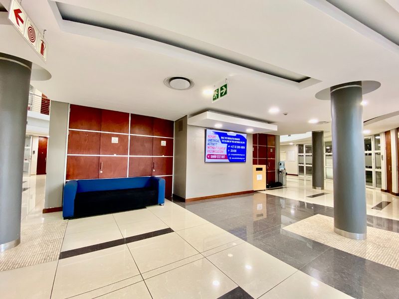 A spacious commercial building lobby featuring polished tiled flooring, structural columns, recessed ceiling lighting, and clear emergency exit signage.