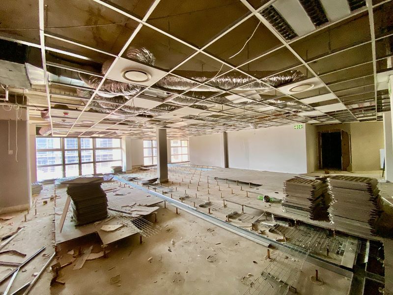 A large, open-plan commercial office space currently undergoing renovation, featuring exposed ceiling infrastructure, raised flooring, and large windows providing natural light.