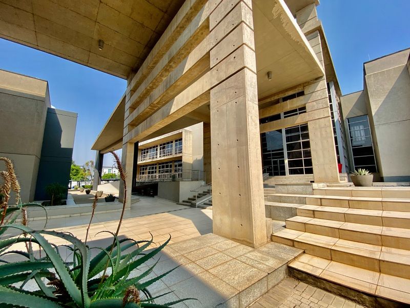Modern commercial building entrance featuring prominent concrete structural columns, a large overhead canopy, and wide tiled stairs leading to a glass-fronted facade.