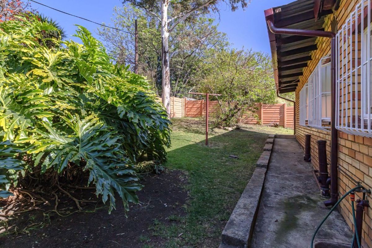 A rear exterior view of the property featuring a brick facade, concrete pathway, and established garden area with perimeter fencing.