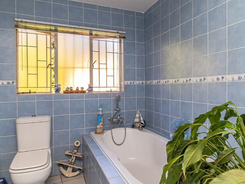 A functional bathroom featuring blue wall tiles, a built-in bathtub, and a standard toilet, with natural light provided by a window with security bars.