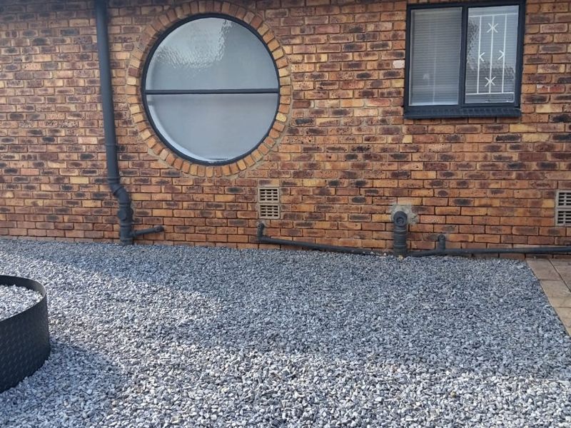 Rear exterior view of a face brick house featuring a distinctive circular window, standard rectangular window with security bars, and a gravel-covered yard area.