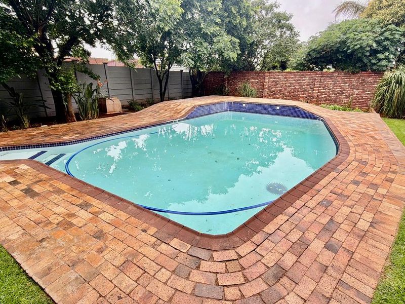 A backyard swimming pool area featuring brick paving, a surrounding lawn, and perimeter boundary walls.