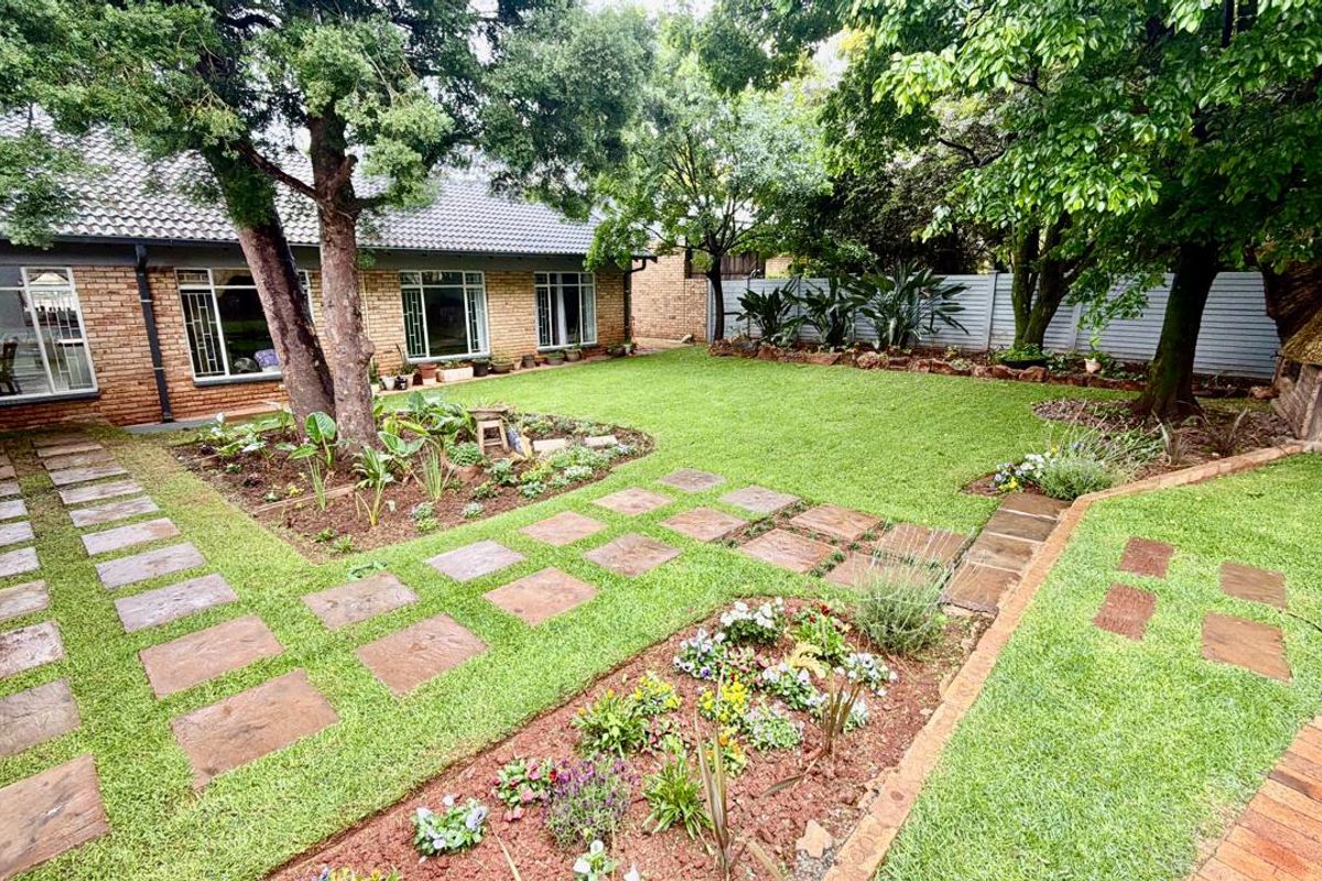 A well-maintained rear garden featuring a brick facade, paved pathways, mature trees, and a built-in brick braai area.