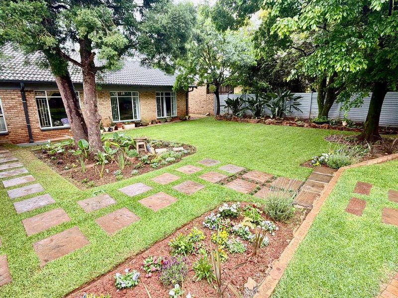 A well-maintained rear garden featuring a brick facade, paved pathways, mature trees, and a built-in brick braai area.
