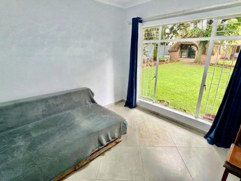 A bright living room featuring large aluminium-framed windows with security bars, tiled flooring, and direct views of the garden.