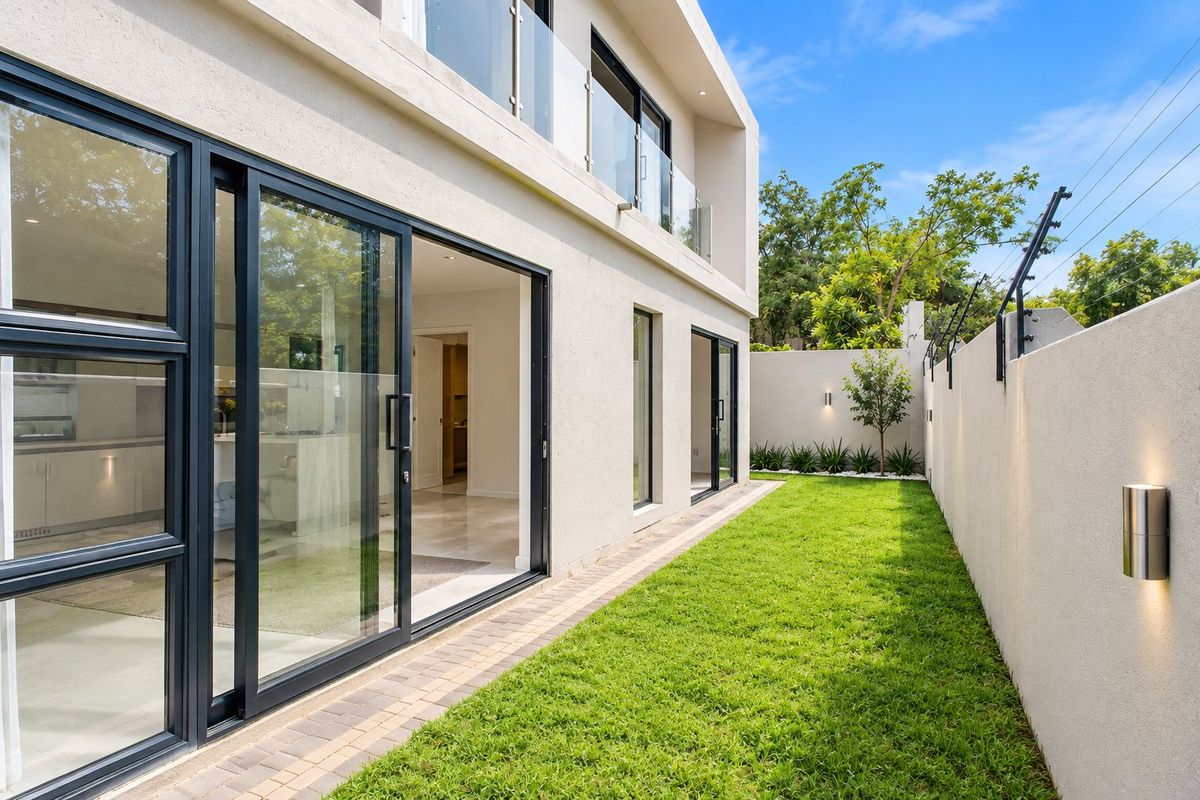 A modern townhouse rear exterior featuring a manicured lawn, paved walkway, sliding glass doors, and a secure boundary wall with integrated lighting.