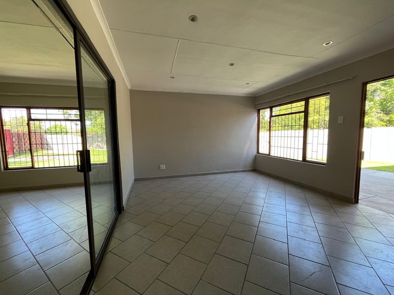 A bright, empty living area featuring neutral tiled flooring, recessed ceiling lighting, and large windows with security bars, offering direct access to the exterior.
