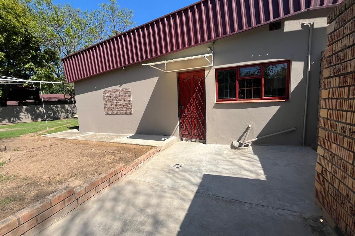 Rear exterior view featuring a concrete patio, security gate, and metal roof structure with adjacent garden space.