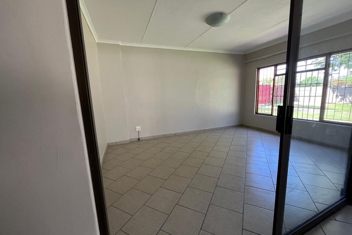 A bright, empty living room featuring neutral tiled flooring, a large window with security bars, and sliding glass door access.