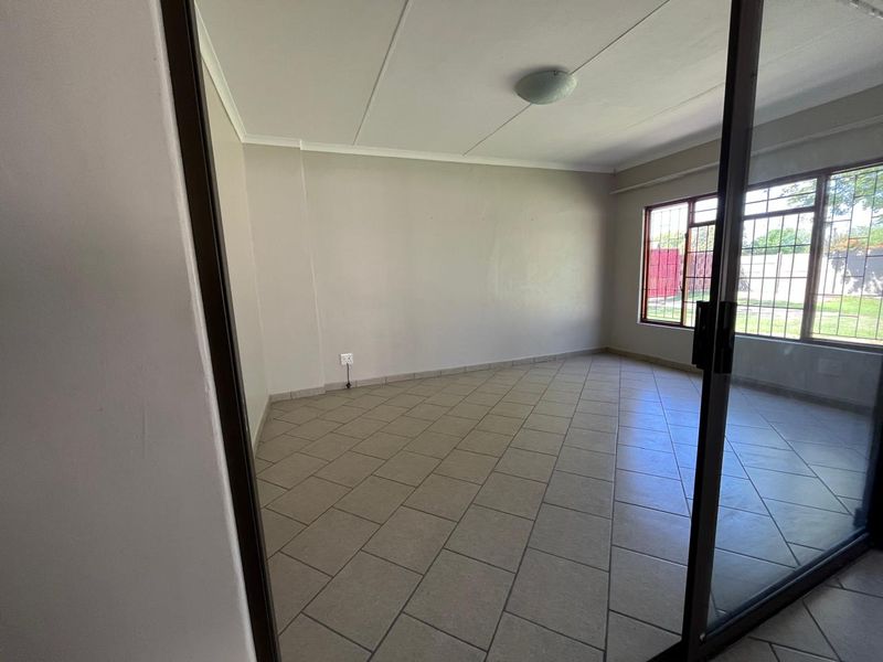 A bright, empty living room featuring neutral tiled flooring, a large window with security bars, and sliding glass door access.