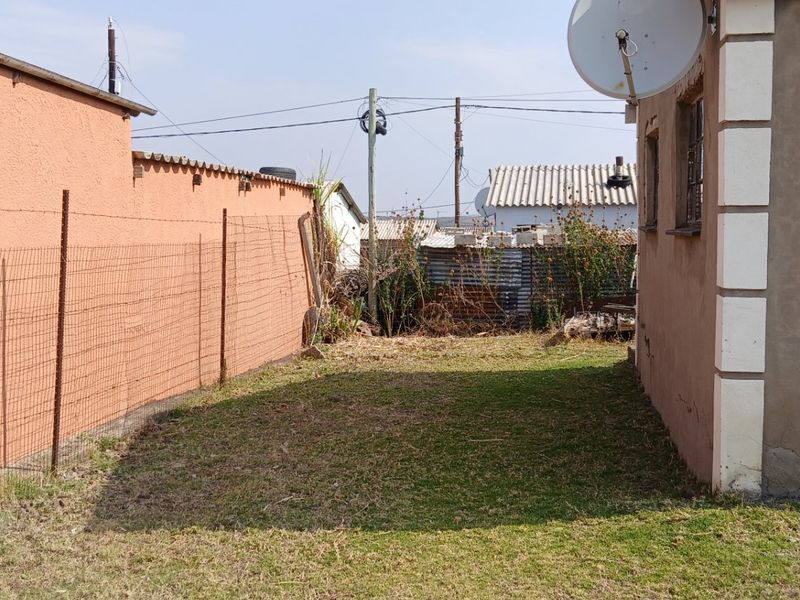 A view of the rear exterior area featuring a lawn, wire mesh fencing, and a plastered boundary wall with a satellite dish mounted on the house.