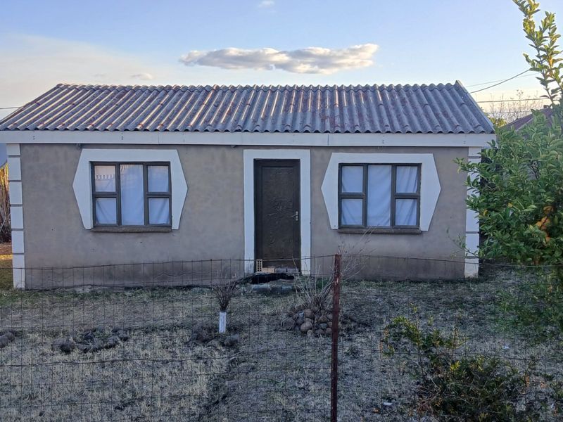 Front elevation of a modest single-story house featuring a corrugated roof, plastered exterior walls with decorative window surrounds, and a wire mesh perimeter fence.