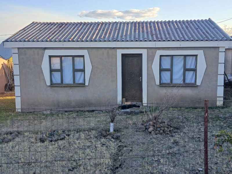Front view of a modest single-story house featuring a corrugated roof, metal-framed windows, and a secure entrance door, set within a fenced yard.