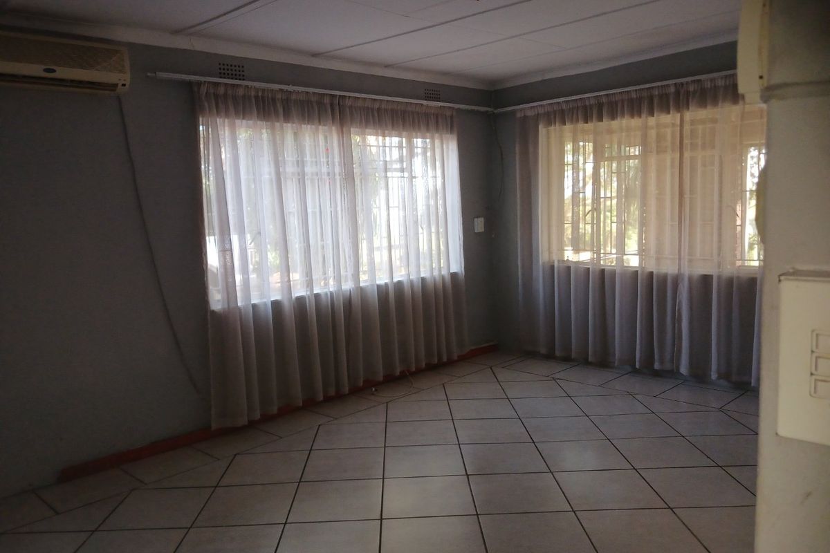 An empty living room featuring tiled flooring, a wall-mounted air conditioning unit, and large windows fitted with security bars and curtain tracks.