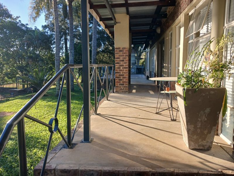 A covered outdoor patio featuring concrete flooring, brick support pillars, and a metal safety railing, overlooking a garden area.