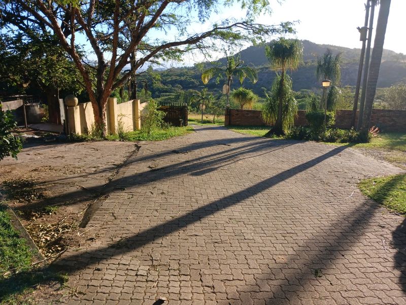 A paved driveway leading towards a gated entrance, featuring mature trees, boundary walls, and scenic mountain views in the background.