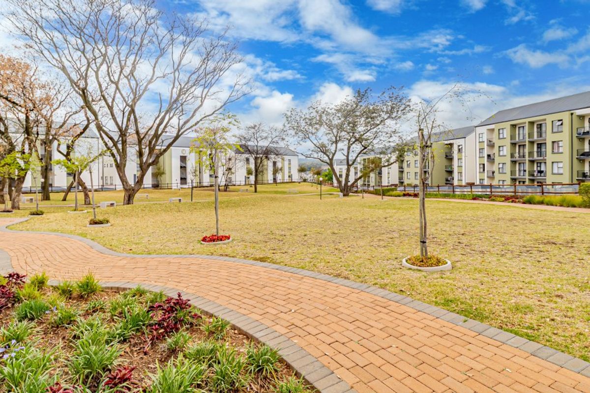 A well-maintained communal garden area featuring a winding brick-paved walkway, manicured lawn, and mature trees within an apartment complex setting.