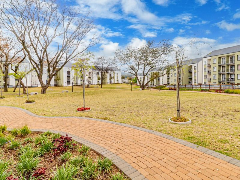 A well-maintained communal garden area featuring a winding brick-paved walkway, manicured lawn, and mature trees within an apartment complex setting.