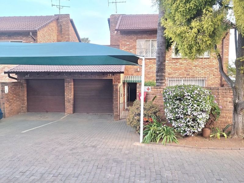 Front exterior view of a brick townhouse featuring a double garage, paved driveway, and a shade cloth carport structure.