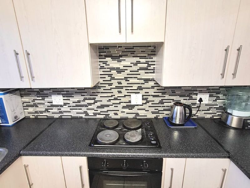 A functional kitchen featuring light wood-finish cabinetry, dark laminate countertops, a mosaic tile backsplash, and a built-in electric oven and hob.