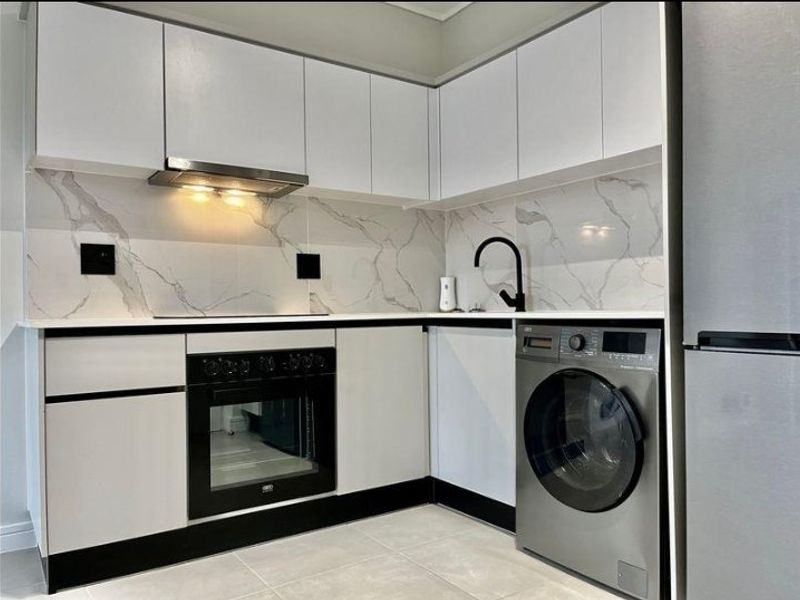 Modern kitchen featuring white cabinetry, marble-look splashback, stone countertops, built-in oven, extractor fan, and integrated laundry appliance space.