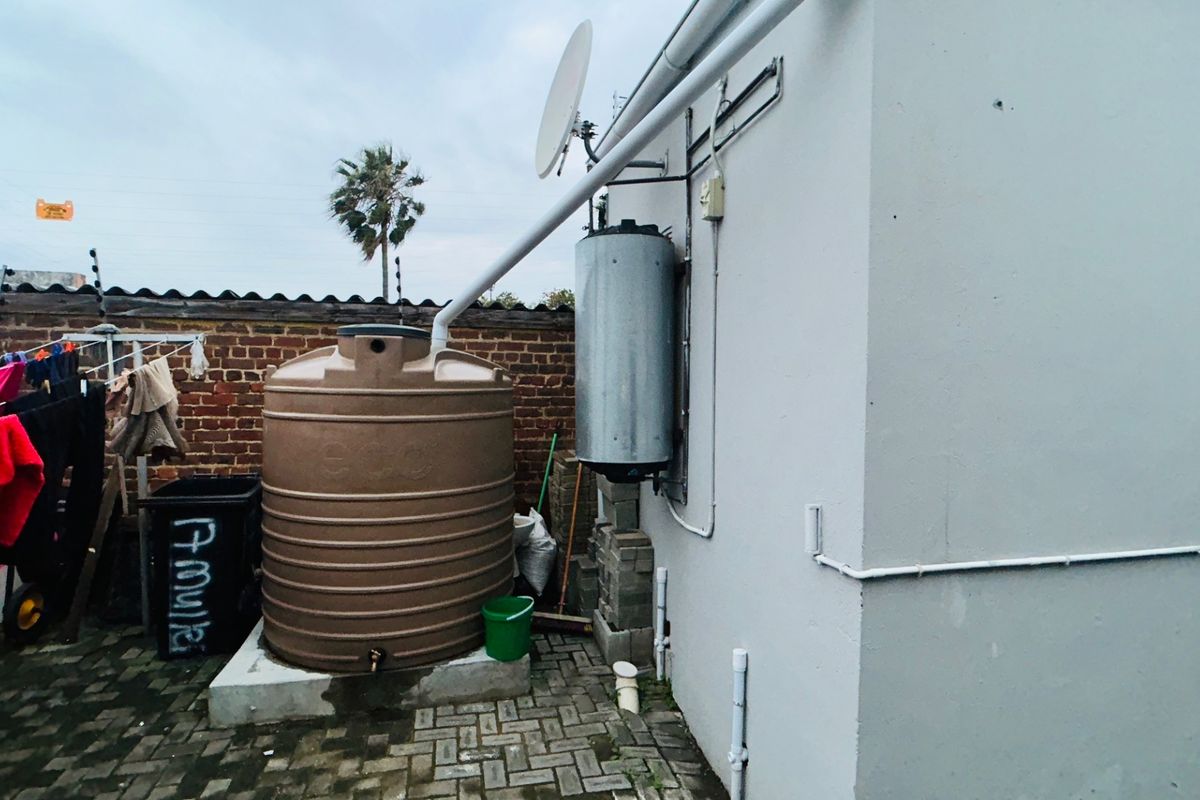 Rear exterior area featuring a large water storage tank, wall-mounted electric geyser, paved ground, and brick boundary wall with electric fencing.