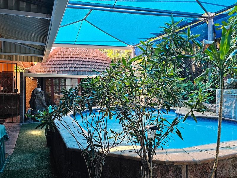 An outdoor pool area featuring a raised swimming pool, stone cladding, and a blue shade sail covering the patio and pool space.