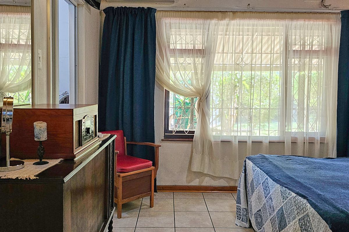 A bedroom featuring tiled flooring and large windows with security bars, offering natural light and a view of the garden.