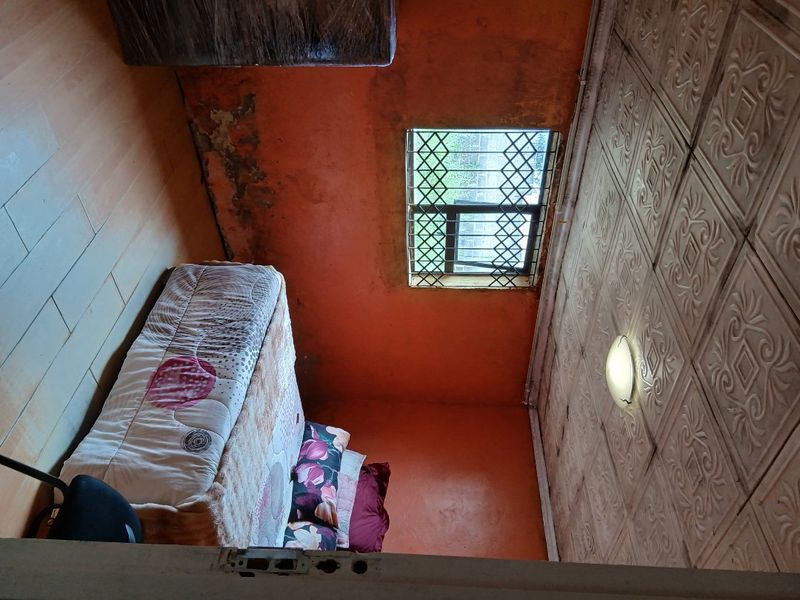 A bedroom featuring decorative ceiling tiles and a security-gated window, currently showing significant signs of wall surface deterioration and peeling paint.