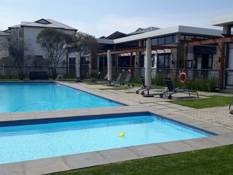 A well-maintained communal swimming pool area featuring paved decking, a timber pergola, and landscaped lawn surrounds within a residential apartment complex.