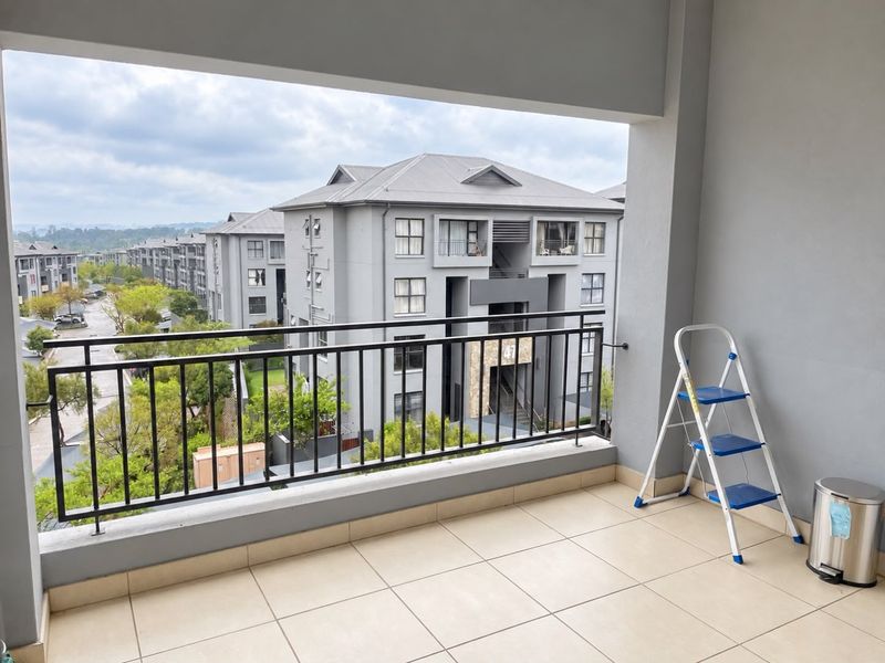 A spacious covered balcony featuring tiled flooring and a metal balustrade, offering an elevated view of the surrounding apartment complex.