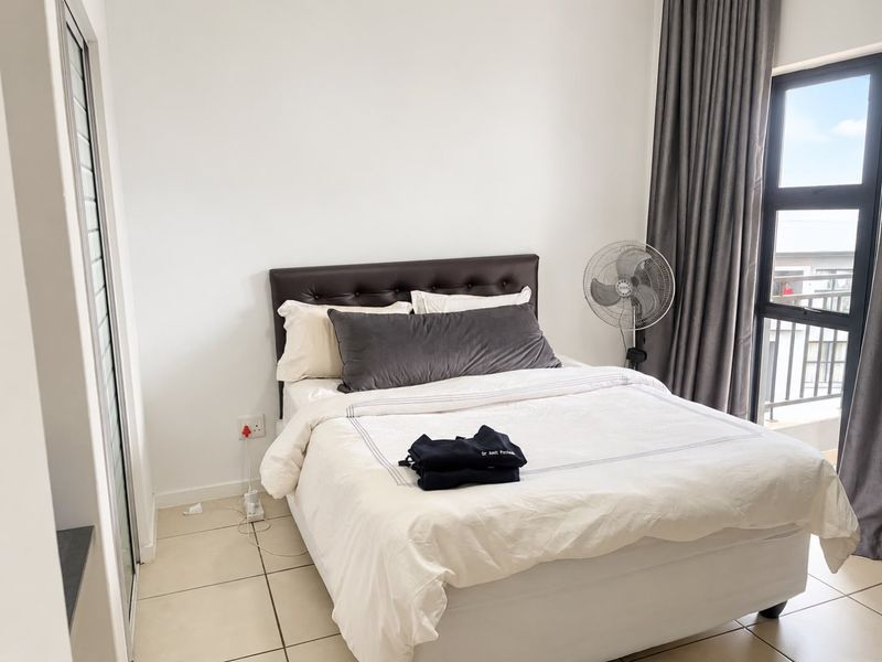 A well-lit bedroom featuring neutral tiled flooring, a built-in cupboard, and a large window with access to a balcony.
