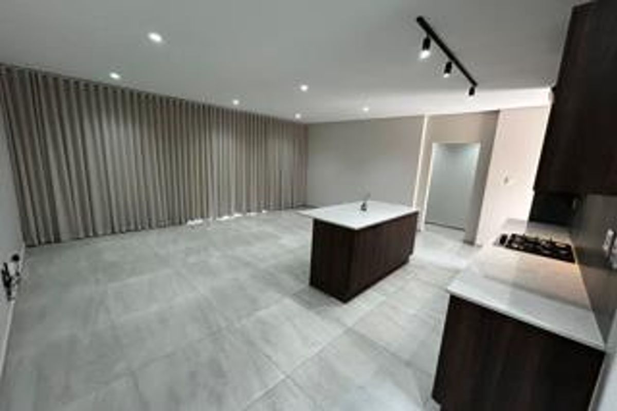 A modern, spacious open-plan living and kitchen area featuring sleek dark cabinetry, a central island with a hob, and neutral tiled flooring.