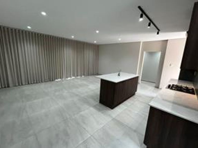 A modern, spacious open-plan living and kitchen area featuring sleek dark cabinetry, a central island with a hob, and neutral tiled flooring.