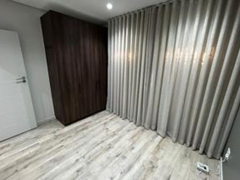 A clean, modern bedroom featuring light-toned laminate flooring, a dark wood built-in wardrobe, and floor-to-ceiling curtains.