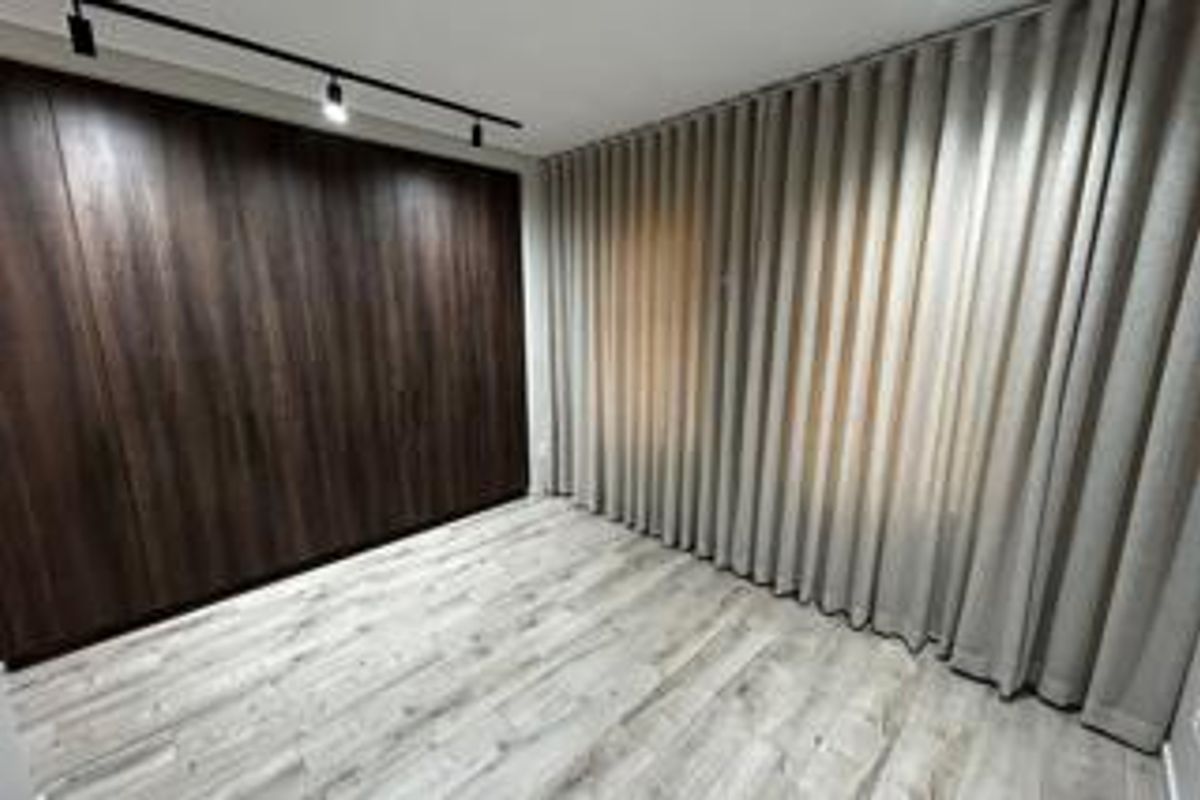 A modern, empty bedroom featuring dark wood-grain built-in cabinetry, light-toned laminate flooring, and full-length window curtains with track lighting.