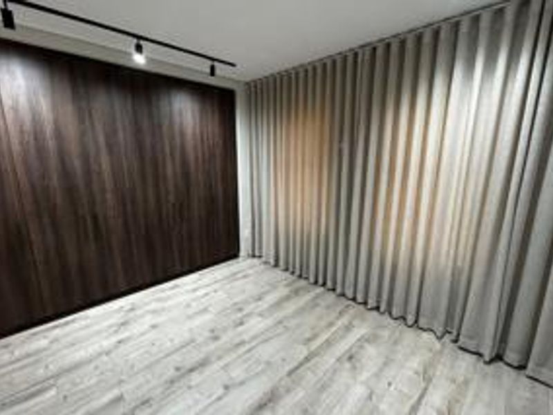 A modern, empty bedroom featuring dark wood-grain built-in cabinetry, light-toned laminate flooring, and full-length window curtains with track lighting.
